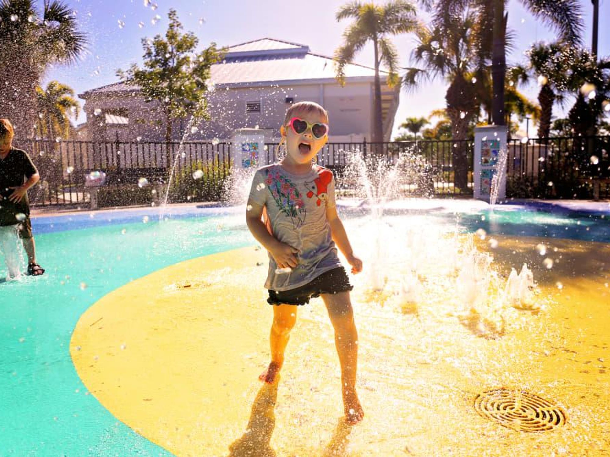 A child in wet clothes and sunglasses stands on a splash pad in the sun.