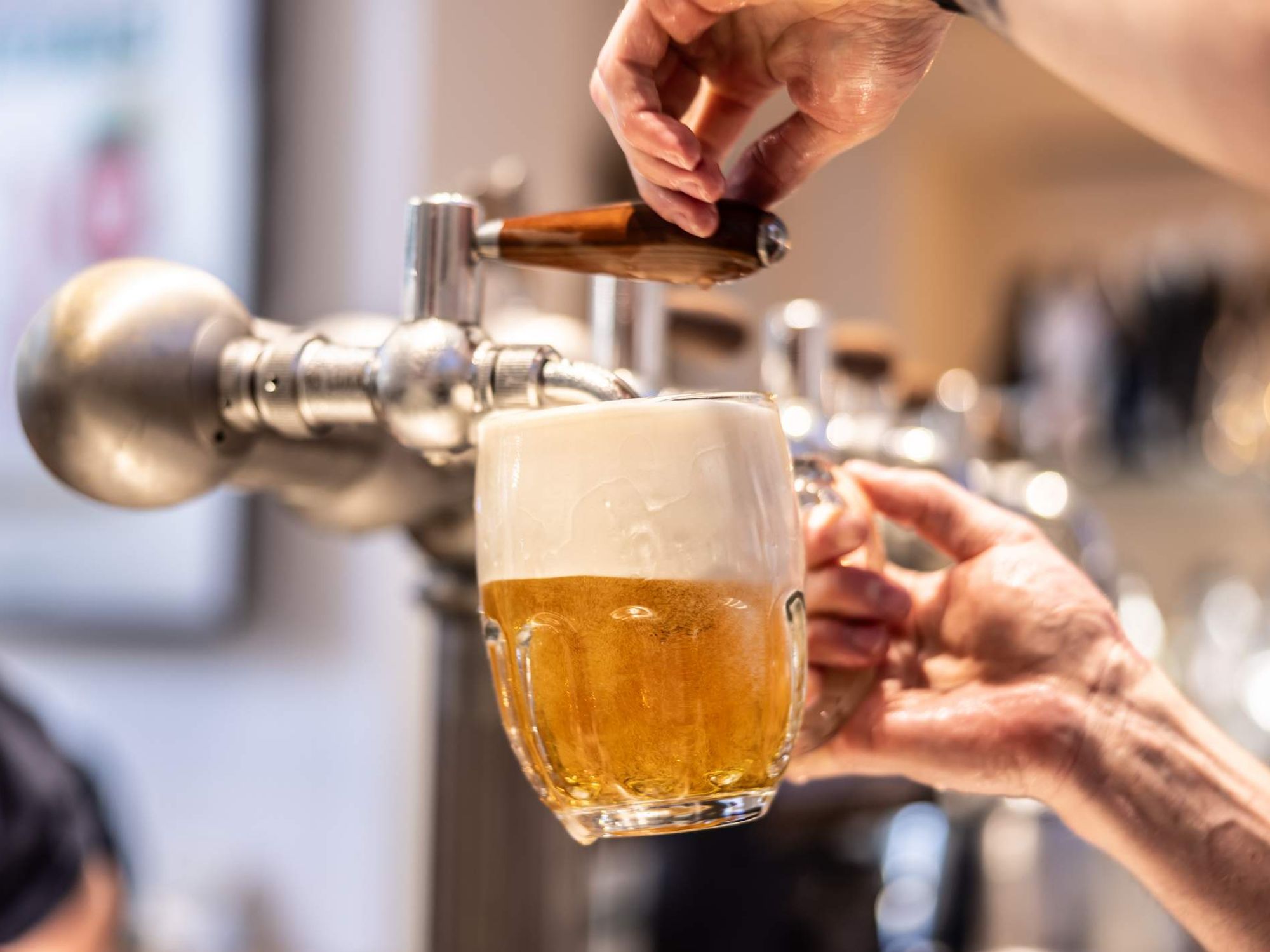 A close-up of an unrecognisable person's hand holding a pint glass at an angle being filled with lager. The lager is golden in colour creating foam and bubbles.