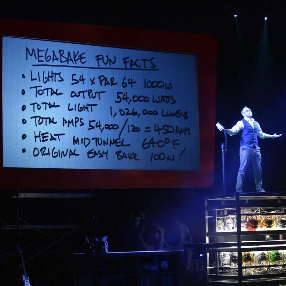 Alton Brown stands on top of a metal rack onstage in front of oven notes.