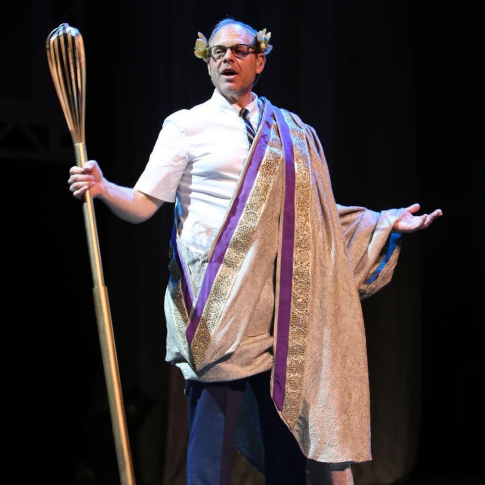 Celebrity chef and TV host Alton Brown poses onstage in a toga with a giant whisk.